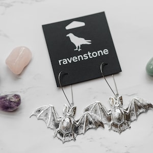 May include: Silver bat earrings with a black card that says "ravenstone" and a white raven logo. The card is on a white marble surface with four small stones.