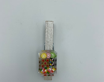 May include: A wooden clothespin with a clear plastic container filled with colorful candies and sweets. The candies include swirls, squares, and round shapes.