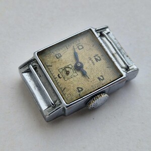 May include: A vintage silver-toned wristwatch with a rectangular face. The watch face is a light gold color with black numerals and hands. The watch has a textured pattern on the face and a small crown on the side.