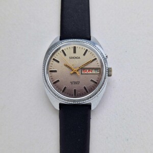 Puede incluir: Un reloj Sekonda vintage con caja plateada y correa de cuero negro. La esfera dorada tiene marcadores y agujas negras, y muestra el día y la fecha. El texto "Sekonda" y "Automatic 25 Jewels" son visibles.