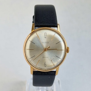 Puede incluir: Un reloj de pulsera dorado vintage con esfera plateada y correa de cuero negro. La esfera del reloj muestra la marca "Wostok", el número 12 y el texto "18 jewels". Un reloj clásico.