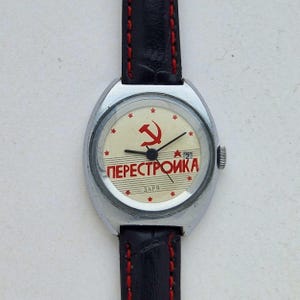 May include: A vintage wristwatch with a silver case and a black leather band with red stitching. The watch face is cream-colored with a red hammer and sickle symbol, the word "PERESTROIKA", and small stars.