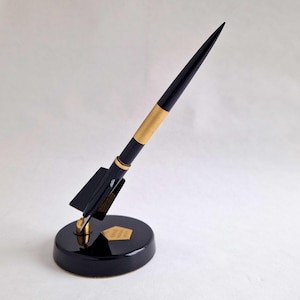Peut inclure: Stylo de bureau en forme de fusée noire et dorée. Le stylo a un corps noir avec une bande dorée et une pointe. Il repose sur une base circulaire noire avec un emblème doré. La fusée est inclinée vers le haut.