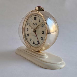 May include: A vintage silver-toned alarm clock with a clear, rounded cover. The clock face is white with black numerals and hands. The clock is marked "SLAVA" and sits on a cream-colored base.