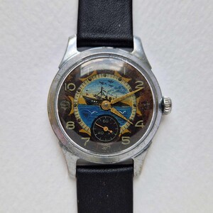 Puede incluir: Un reloj de pulsera plateado con correa de cuero negro. La esfera presenta una escena pintada de un barco en el mar con un motivo de rayos de sol, manecillas doradas y una pequeña subesfera negra en la parte inferior. Los números están dispuestos en círculo.
