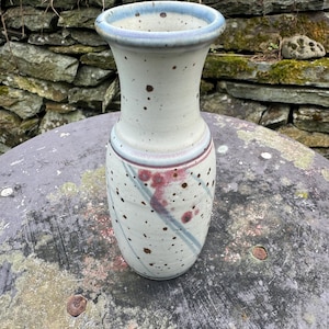 May include: A ceramic vase with a flared rim, featuring a speckled off-white glaze, blue-tinted edges, and diagonal bands of muted pink and grey accents. The body of the vase has a cylindrical neck that widens into a rounded base.