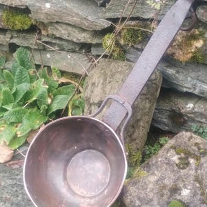 May include: A vintage copper pot with a long iron handle. The pot is round and has a smooth, worn surface. The handle is attached to the pot with two metal rings.