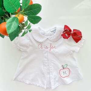 K&ouml;nnte beinhalten: Wei&szlig;es Baby-Shirt mit einem roten gestickten Apfel und dem Text "A is for".