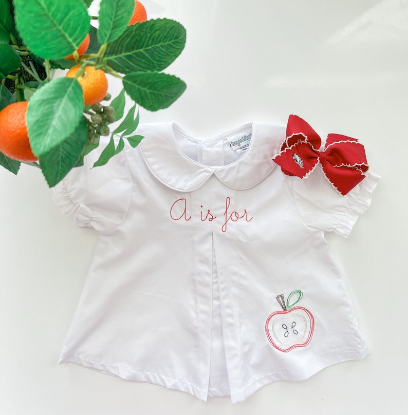 K&ouml;nnte beinhalten: Wei&szlig;es Baby-Shirt mit einem roten gestickten Apfel und dem Text "A is for".