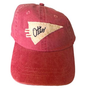 May include: A red baseball cap with a curved brim. The cap features a white pennant-shaped patch with the name "Otto" embroidered in black script. The patch also includes red and white stripes. The cap is made of a soft, worn material.