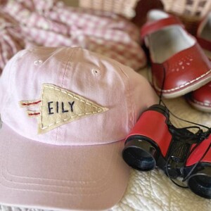 以下が含まれることがあります： ベージュのペナントに「EILY」と書かれた薄いピンク色の野球帽、赤い双眼鏡、赤い革製のメリージェーンシューズのペア。ピンクと白の模様の生地が背景にあります。