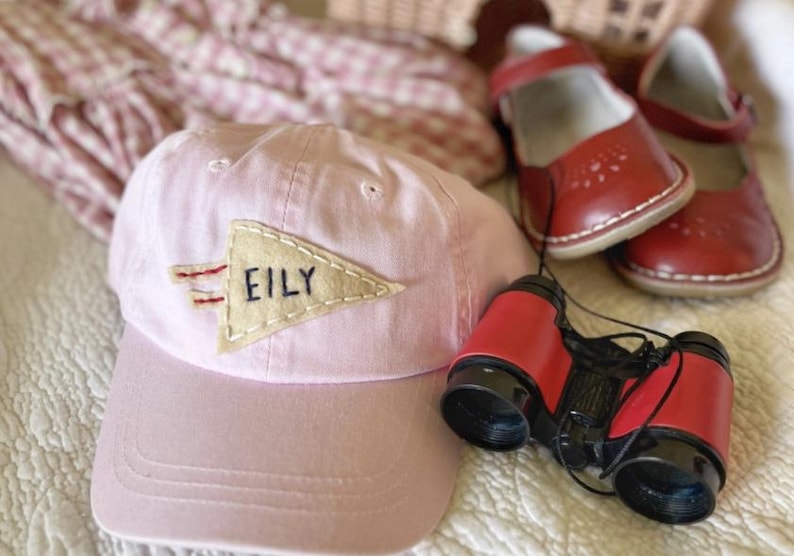 以下が含まれることがあります： ベージュのペナントに「EILY」と書かれた薄いピンク色の野球帽、赤い双眼鏡、赤い革製のメリージェーンシューズのペア。ピンクと白の模様の生地が背景にあります。