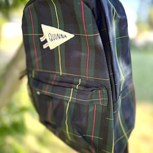 May include: A backpack featuring a dark green, navy blue, yellow, and red tartan pattern. The front of the bag displays a white triangular pennant patch with the name QUINNA embroidered in black thread. It includes a zippered front pocket.