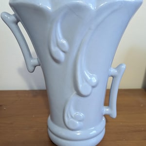 May include: A light blue ceramic vase with a flared top and two curved handles. The vase features embossed floral designs and stands approximately 8 inches tall.