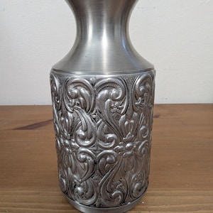 May include: A silver-toned metal vase featuring a smooth, flared neck and a cylindrical body decorated with an intricate, raised floral and scrollwork pattern. The metallic surface has a polished finish with darker recessed areas in the relief.