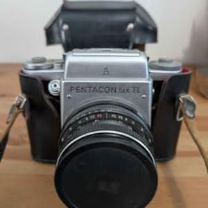 May include: A silver Pentacon Six TL camera with a black lens and leather case. The camera has the text "PENTACON six TL" on the front. The lens is covered with a black lens cap. The camera is on a wooden surface.