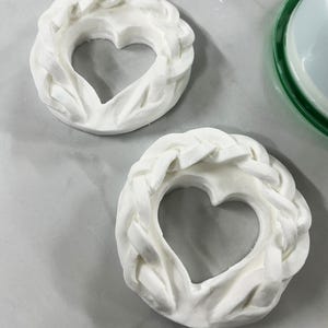 Puede incluir: Dos piezas decorativas blancas en forma de corazón. Cada pieza tiene un borde trenzado y un recorte en forma de corazón en el centro. Las piezas están sobre una superficie gris claro. El diseño es simple y elegante.
