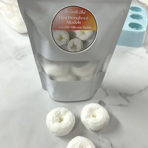 Puede incluir: Un paquete de mini modelos de donuts para moldes de silicona DIY. El paquete tiene una pegatina circular con el texto "The Candle Chef Mini Doughnut Models For DIY Silicone Molds". Tres mini modelos de donuts blancos están delante del paquete.