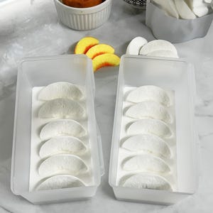 Puede incluir: Dos recipientes de plástico transparente llenos de golosinas blancas en forma de media luna, probablemente malvaviscos o caramelos. Los recipientes están sobre una superficie clara, con golosinas adicionales y un cuenco pequeño de rollos de canela en el fondo. Tema: panadería o postre.