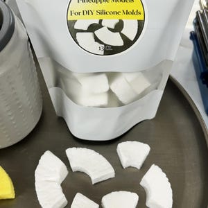 Puede incluir: Una bolsa blanca etiquetada "The Candle Chef" contiene modelos de piña para moldes de silicona DIY. Varios modelos blancos con forma de piña están dispuestos en una bandeja, junto a un objeto amarillo. La bolsa está marcada "13 Ct."