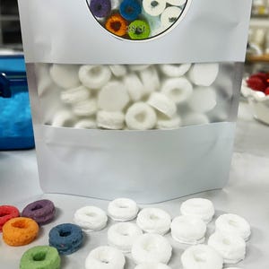 Puede incluir: Una bolsa de modelos de cera blancos en forma de rosquilla para moldes de silicona DIY. La bolsa tiene una ventana transparente y una etiqueta que dice "Large Fruity Loop Models". Varios modelos de rosquillas en miniatura de colores están esparcidos alrededor de la bolsa.