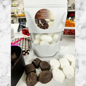 Puede incluir: Una bolsa blanca con una ventana transparente que muestra modelos de caramelos blancos. La bolsa tiene el texto "The Candle Chef Chocolate Candy Models For DIY Silicone Molds". Varios caramelos de chocolate y blancos están dispuestos sobre una superficie blanca.