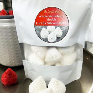 Puede incluir: Una bolsa blanca etiquetada "The Candle Chef" que contiene modelos de fresas enteras para moldes de silicona DIY. La bolsa tiene una ventana transparente que muestra los modelos de fresas blancas. Algunos modelos de fresas blancas están fuera de la bolsa en una bandeja plateada. También se ve una fresa roja.