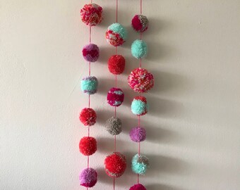May include: A wooden hoop with multiple strands of yarn hanging from it. Each strand has a series of colourful pom-poms in shades of pink, red, grey, and turquoise.