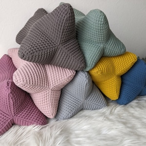 Könnte beinhalten: Ein Stapel von sechs sternförmigen Kissen in verschiedenen Farben, darunter Grau, Grün, Gelb, Rosa, Blau und Hellgrau. Die Kissen sind aus einem Waffelstoff gefertigt.