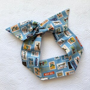 May include: A blue headband with a vintage postage stamp print. The headband is tied in a bow and features stamps from various countries, including Spain, Egypt, and Sydney. The text "PAR AVION" is printed on the headband.