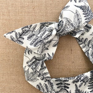May include: A white fabric headband with a black fern pattern. The headband is tied in a bow.