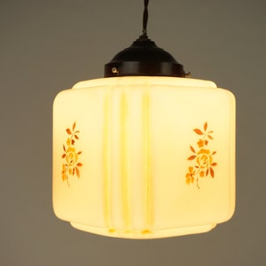 May include: A white glass pendant light fitting with a black metal ceiling rose. The glass shade is square shaped with rounded edges and features a floral design in shades of brown and yellow.