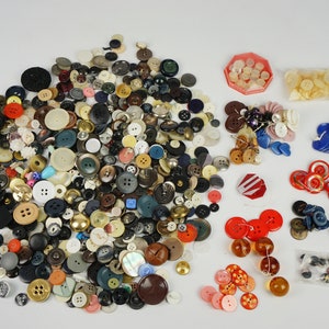 Op de afbeelding: Een verzameling diverse knopen in verschillende maten, vormen en kleuren. De knopen zijn verspreid over een wit oppervlak, sommige gegroepeerd in kleine bakjes. Kleuren zijn zwart, wit, bruin, rood en blauw.