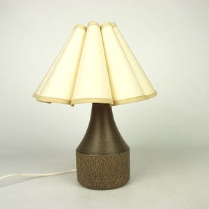 Może przedstawiać: Brązowa ceramiczna lampa stołowa z plisowanym białym abażurem z tkaniny. Lampa ma teksturowaną podstawę.