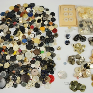 Puede incluir: Una gran colección de botones surtidos en varios colores, formas y tamaños. Los botones están hechos de diferentes materiales, incluyendo plástico, metal y madera. Algunos de los botones tienen diseños decorativos, mientras que otros son lisos. Los botones están dispuestos en una pila sobre una superficie blanca.