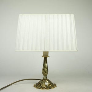 Puede incluir: Una lámpara de mesa dorada con una pantalla de lámpara blanca plisada. La lámpara tiene una base decorativa y un diseño sencillo.