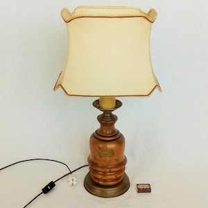 Peut inclure: Une lampe de table en bois avec un abat-jour en tissu beige. La lampe a une base en laiton et un corps en bois avec une finition texturée. La lampe est sur une surface blanche.