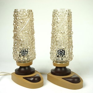 Może przedstawiać: Para lamp stołowych w stylu vintage. Każda lampa ma przezroczysty, teksturowany szklany klosz z wzorem bąbelków. Klosze osadzone są na brązowej podstawie z beżową, łezkowatą stopą. Lampy podłączone są do białego przewodu zasilającego.