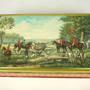May include: A vintage tin box featuring a colourful illustration of a hunting scene. The scene shows riders on horseback with hounds in a wooded area. The box has a gold trim and a red and white patterned base.