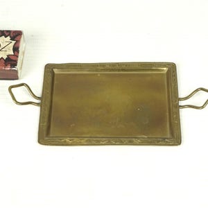 Könnte beinhalten: Ein kleines, rechteckiges Tablett aus Messing mit zwei Griffen. Das Tablett hat einen dekorativen Rand und eine glatte, polierte Oberfläche.