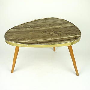 Op de afbeelding: Een vintage salontafel met een uniek, asymmetrisch ontwerp. Het tafelblad heeft een bruin en goud gemarmerd patroon, omlijst door een goudkleurige metalen rand. Hij staat op drie schuine, lichtgekleurde houten poten. De tafel is ongeveer 75 cm lang.