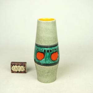 May include: A tall, light green ceramic vase with a yellow interior. The vase features a band of turquoise with two orange circles and black accents. A small box with text is next to the vase.