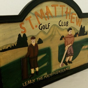 Op de afbeelding: Een houten bord met een zwart frame en een geschilderd tafereel van twee golfers. Het bord heeft de tekst "St. Matthew Golf Club" en "Learn the Magnificence Game".