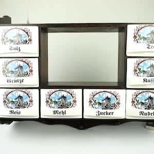 May include: A dark wooden spice rack with eight white ceramic spice containers. Each container has a windmill illustration and a German word. The words are: Salz, Grütze, Reis, Mehl, Zucker, Nudel, Tee, and Kaffee.