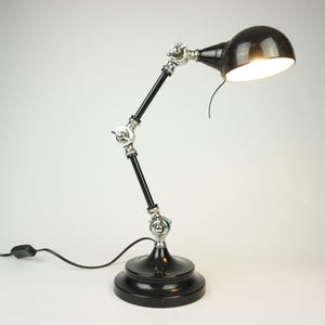 Könnte beinhalten: Eine schwarze Schreibtischlampe mit rundem Schirm und verstellbarem Arm. Die Lampe hat einen kreisförmigen Sockel und silberfarbene Gelenke. Die Lampe ist an eine Steckdose angeschlossen.