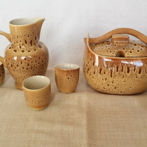May include: A set of brown ceramic serving dishes with a speckled glaze. The set includes a jug, a large bowl with a wooden handle, and six small cups.