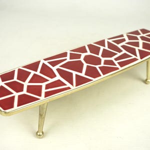 Puede incluir: Una mesa de centro con una tapa de patrón geométrico rojo y blanco y patas de metal dorado.