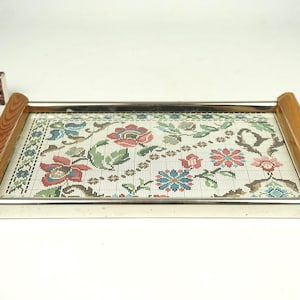 Op de afbeelding: Rechthoekig dienblad met een bloemen tapijtontwerp. De lade heeft een zilveren metalen frame en houten handvatten. Het tapijt heeft rode, blauwe en groene bloemen op een crèmekleurige achtergrond. Een klein doosje lucifers is zichtbaar.