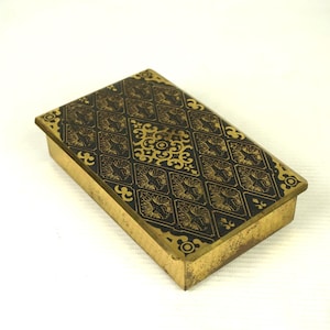 May include: A small, rectangular box with a gold-coloured metal exterior and a black, patterned top. The pattern is intricate and features a repeating design of stylised flowers and leaves.