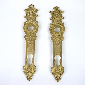 Op de afbeelding: Twee antieke messing deurklinkplaten. Elke plaat heeft sierlijke details, waaronder een gezicht, bloemmotieven en een sleutelgat. De platen zijn goudkleurig en symmetrisch van ontwerp.